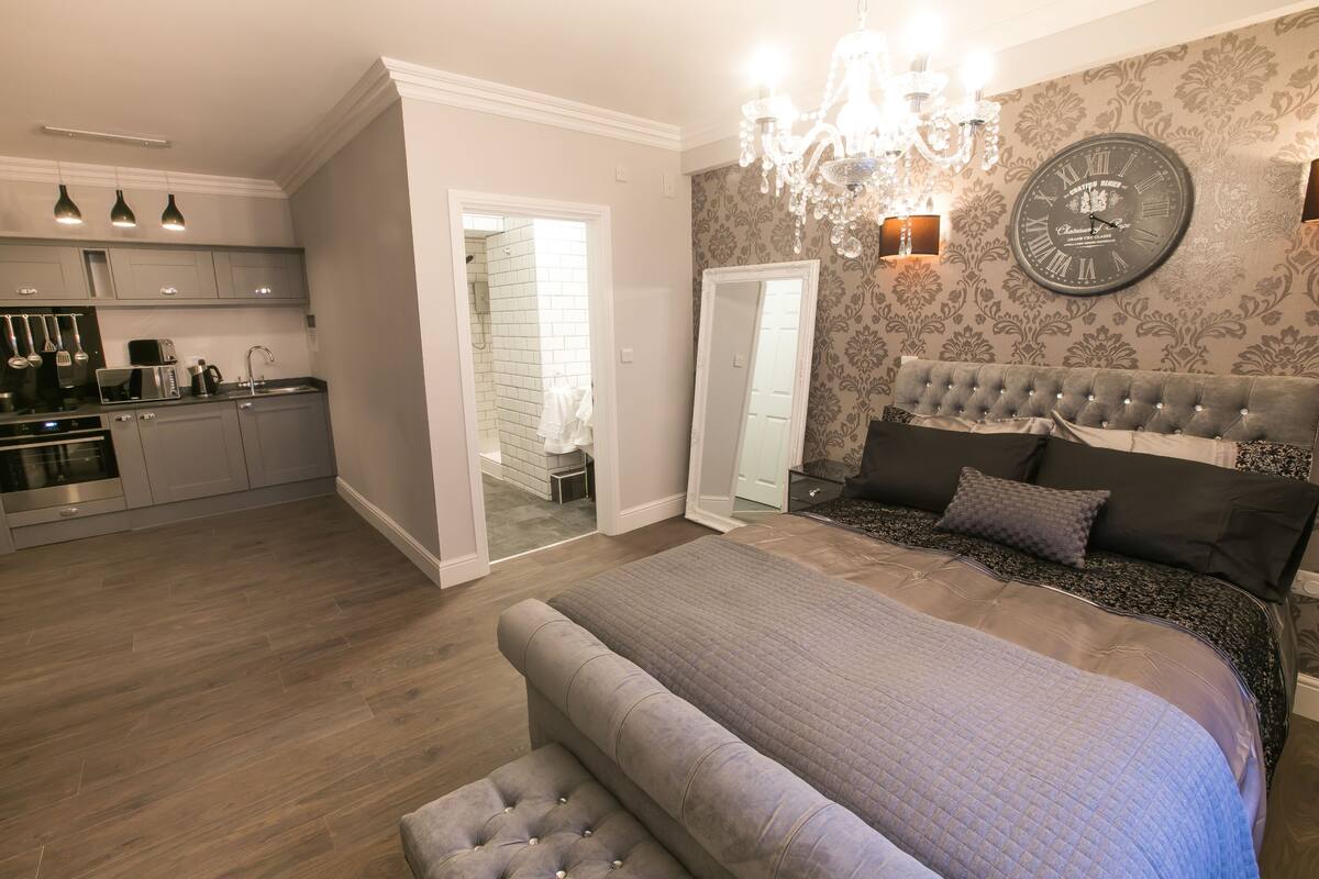 A bedroom with a large bed, a chandelier, a clock, and a view into the kitchen.