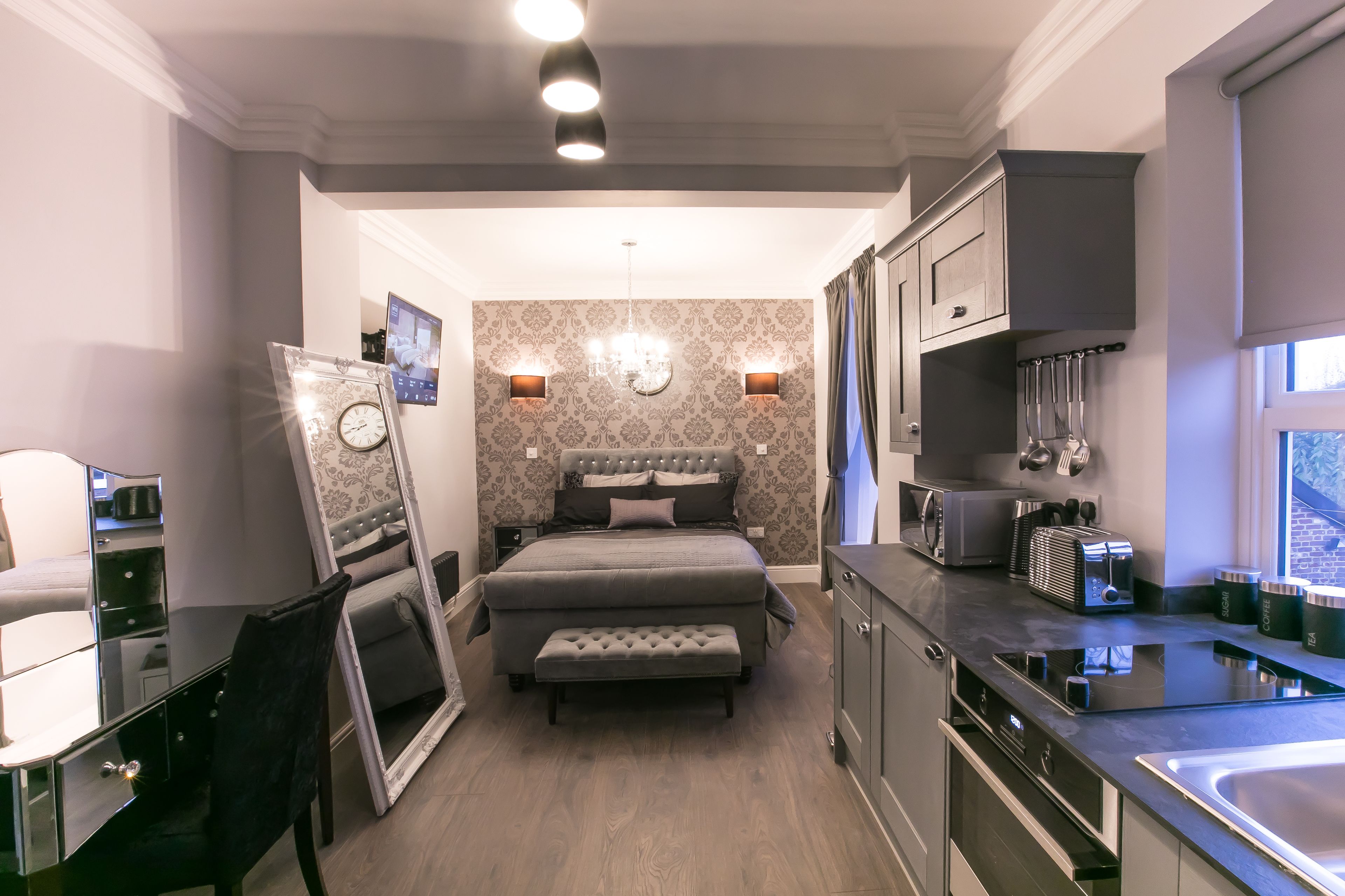 A compact bedroom with a bed, bedside table, and a mirror. The room has a small kitchenette with a sink, stove, and cabinets.