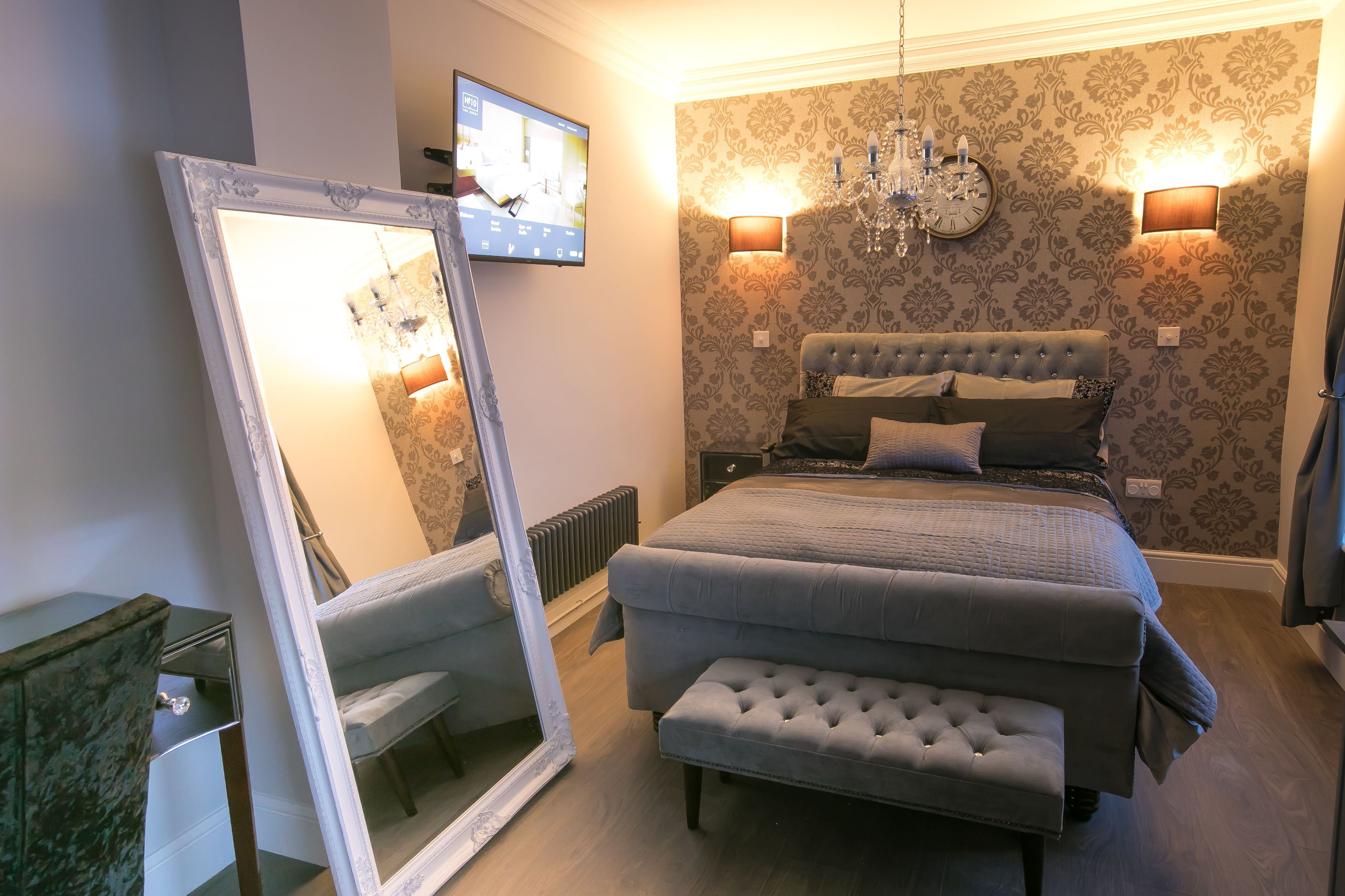 A bedroom with a large bed, a bench, a mirror, a TV, and a chandelier.