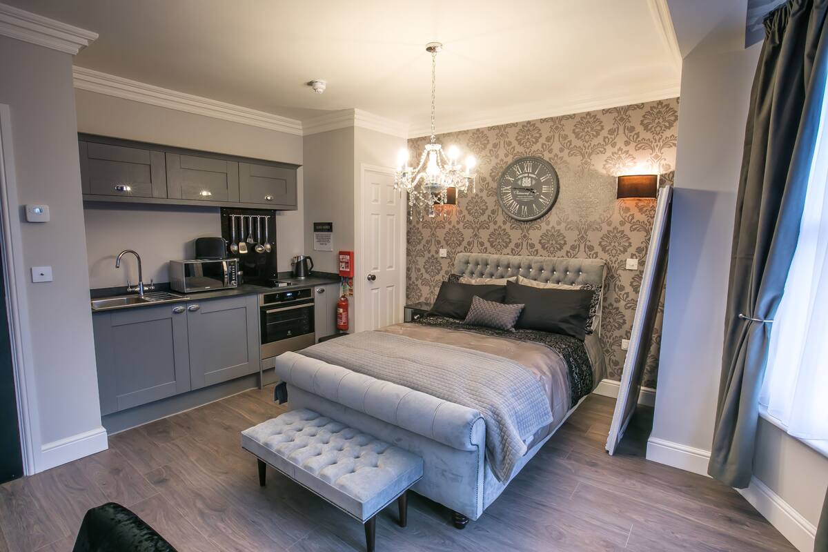 A bedroom with a large bed, a small bench, a chandelier, a clock, and a kitchen area.