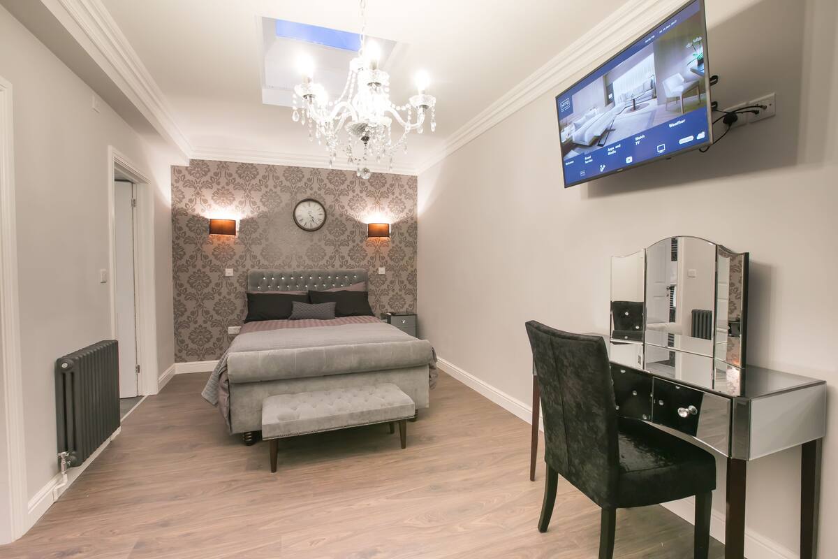 A modern bedroom with a bed, a desk, a chair, a TV, and a chandelier.