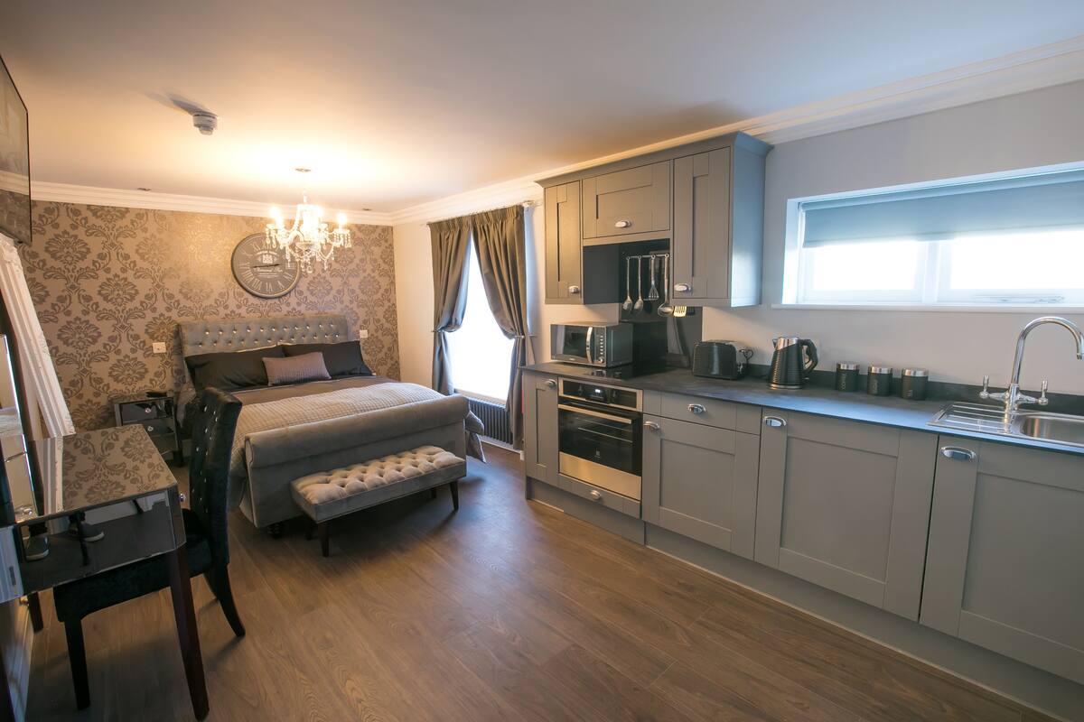 A bedroom with a large bed, a bedside table, and a chair. The room has a wooden floor and a chandelier. There is a window with curtains and a view of the outside.