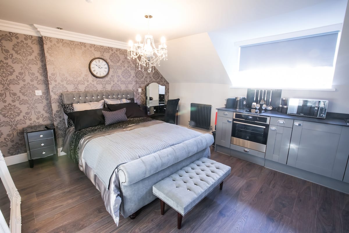 A bedroom with a large bed, a vanity, a chandelier, and a kitchenette.