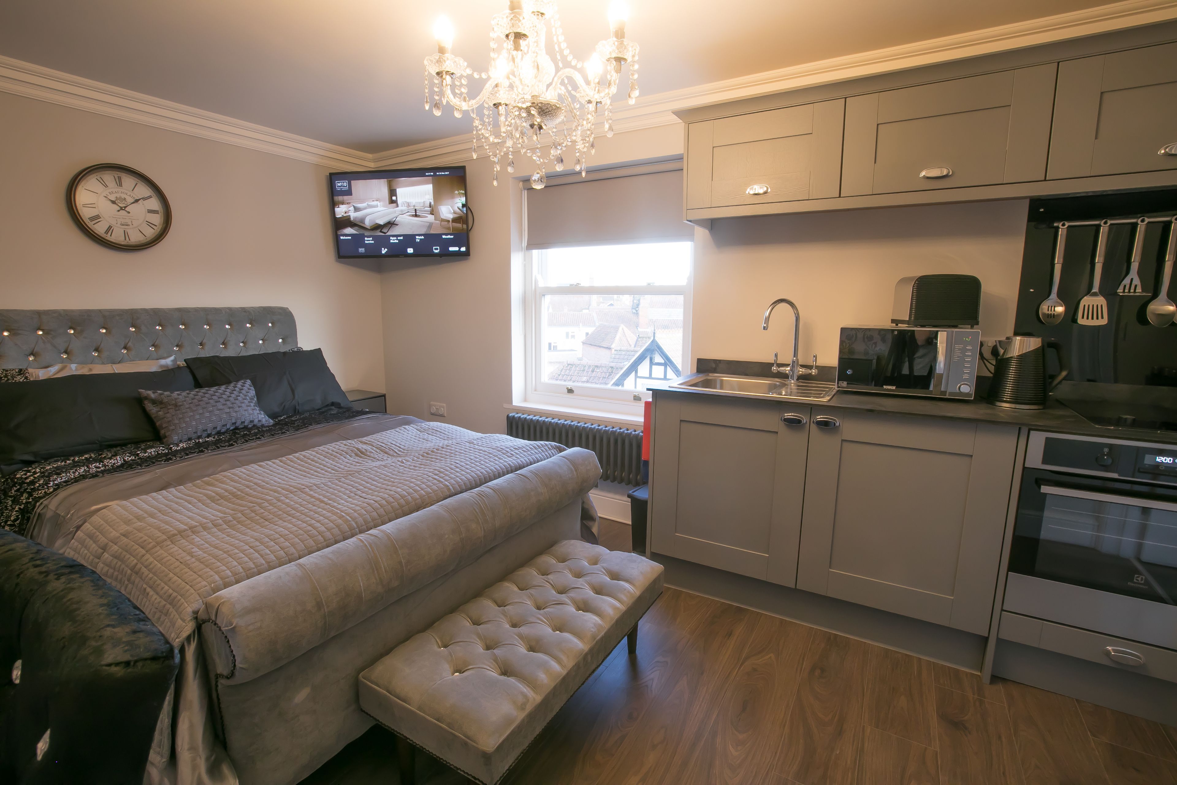 A bedroom with a large bed, a bench, a TV mounted on the wall, and a kitchen area with cabinets and appliances.