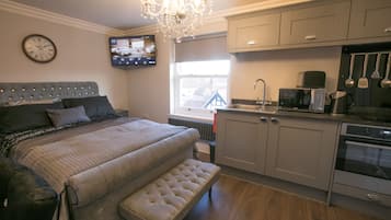 A bedroom with a large bed, a bench, a TV mounted on the wall, and a kitchen area with cabinets and appliances.