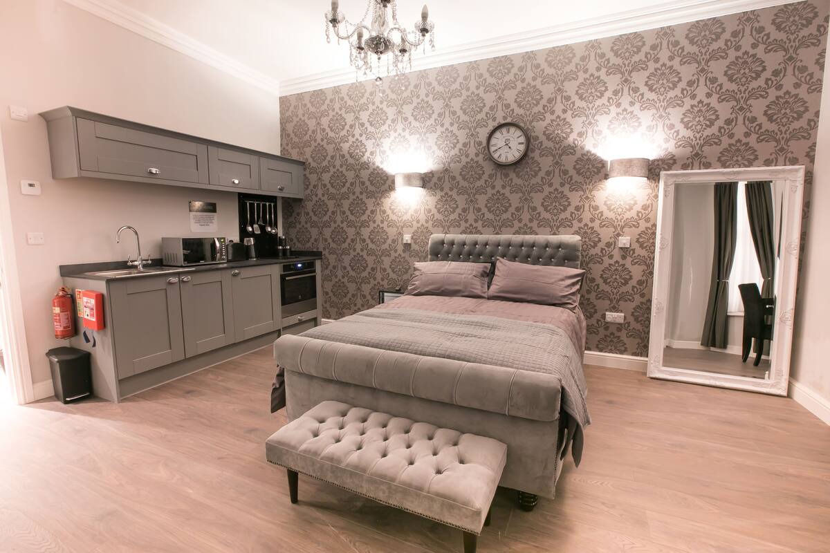 A bedroom with a large bed, a bedside table, a mirror, and a built-in kitchen area with cabinets and appliances.