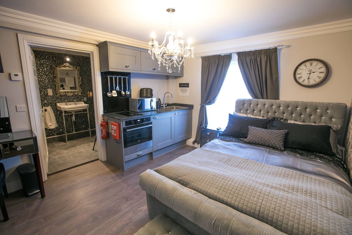 A hotel room with a bed, a small kitchenette, a bathroom, and a chandelier.