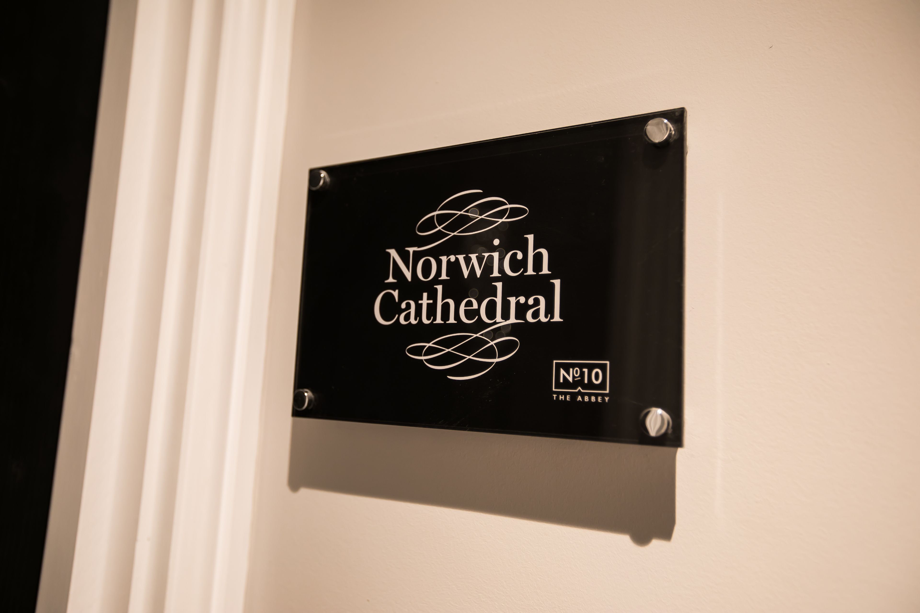 Un senyal negre amb lletres blanques a la paret que indica 'Norwich Cathedral' i 'No.10 The Abbey'.