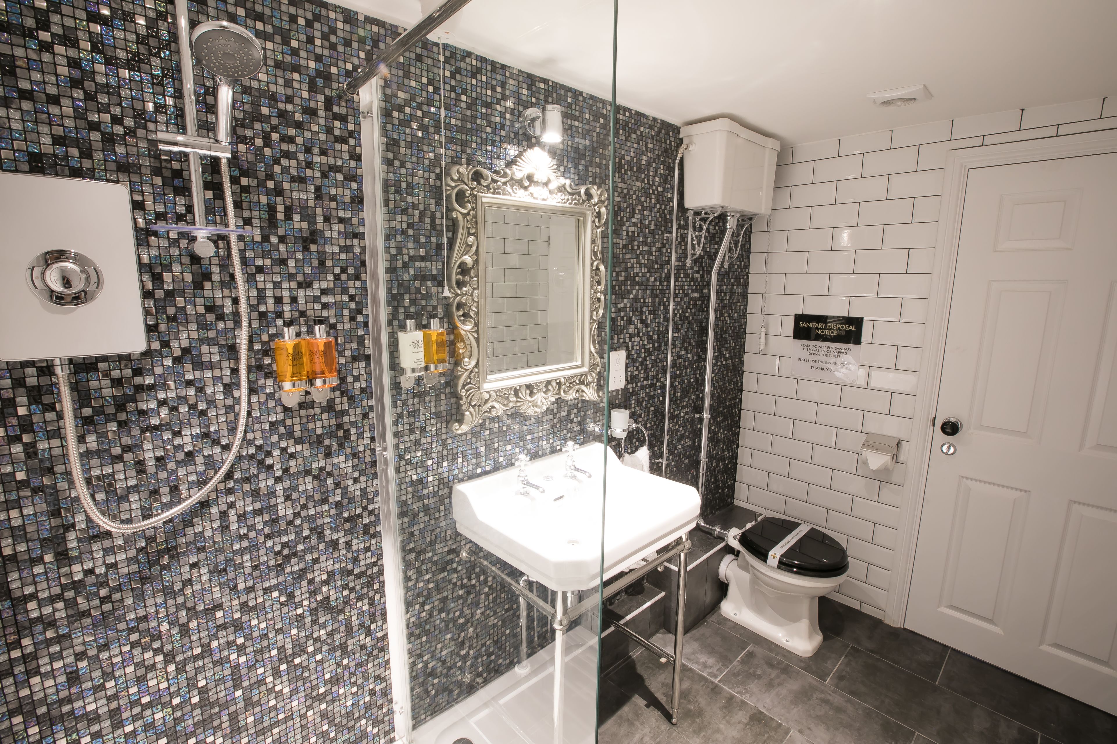 Un baño con ducha, inodoro y lavamanos, con un revestimiento de mosaicos y una puerta blanca.