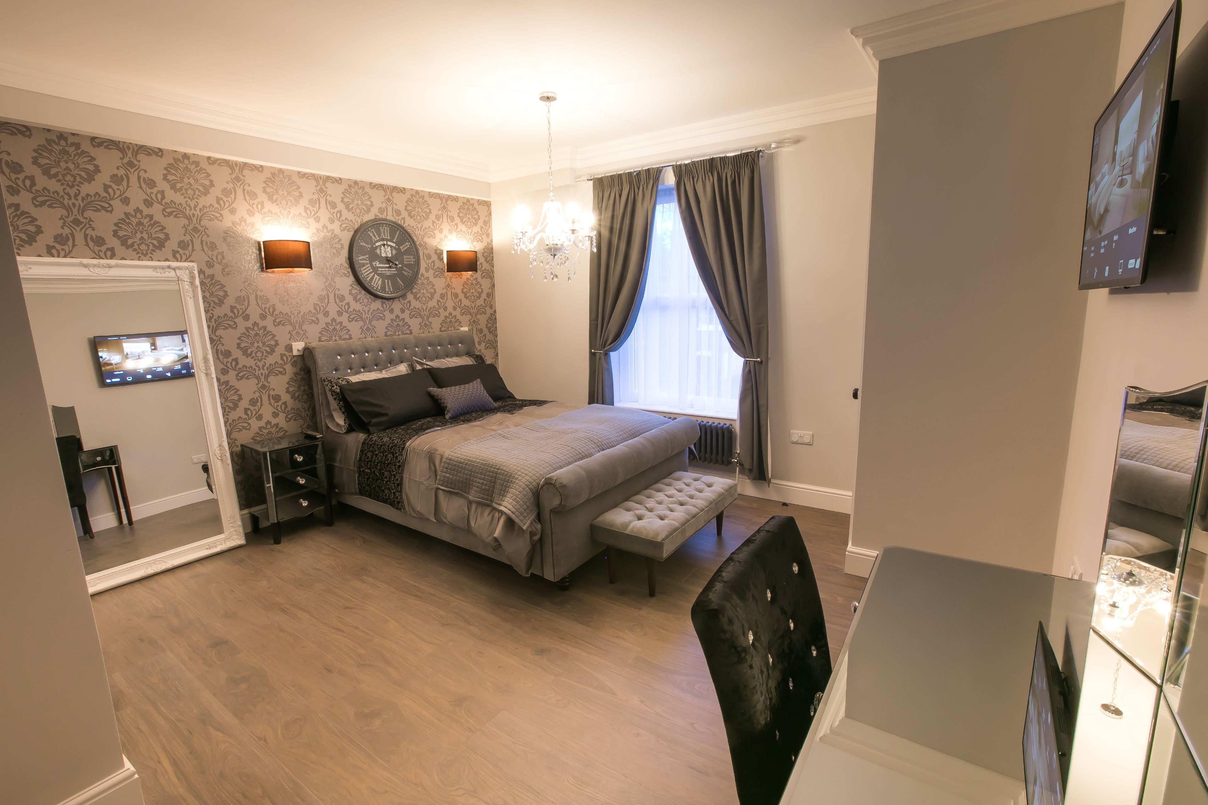 A bedroom with a bed, bedside tables, a mirror, a chandelier, a window with curtains, and a television mounted on the wall.
