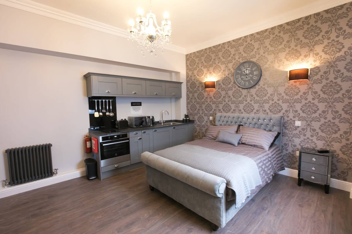 A bedroom with a large bed, a small bedside table, a nightstand, a built-in kitchen with a sink and cabinets, a chandelier, and a clock on the wall.