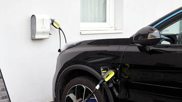 A black car parked next to a white wall with a window and a wall-mounted electrical box.