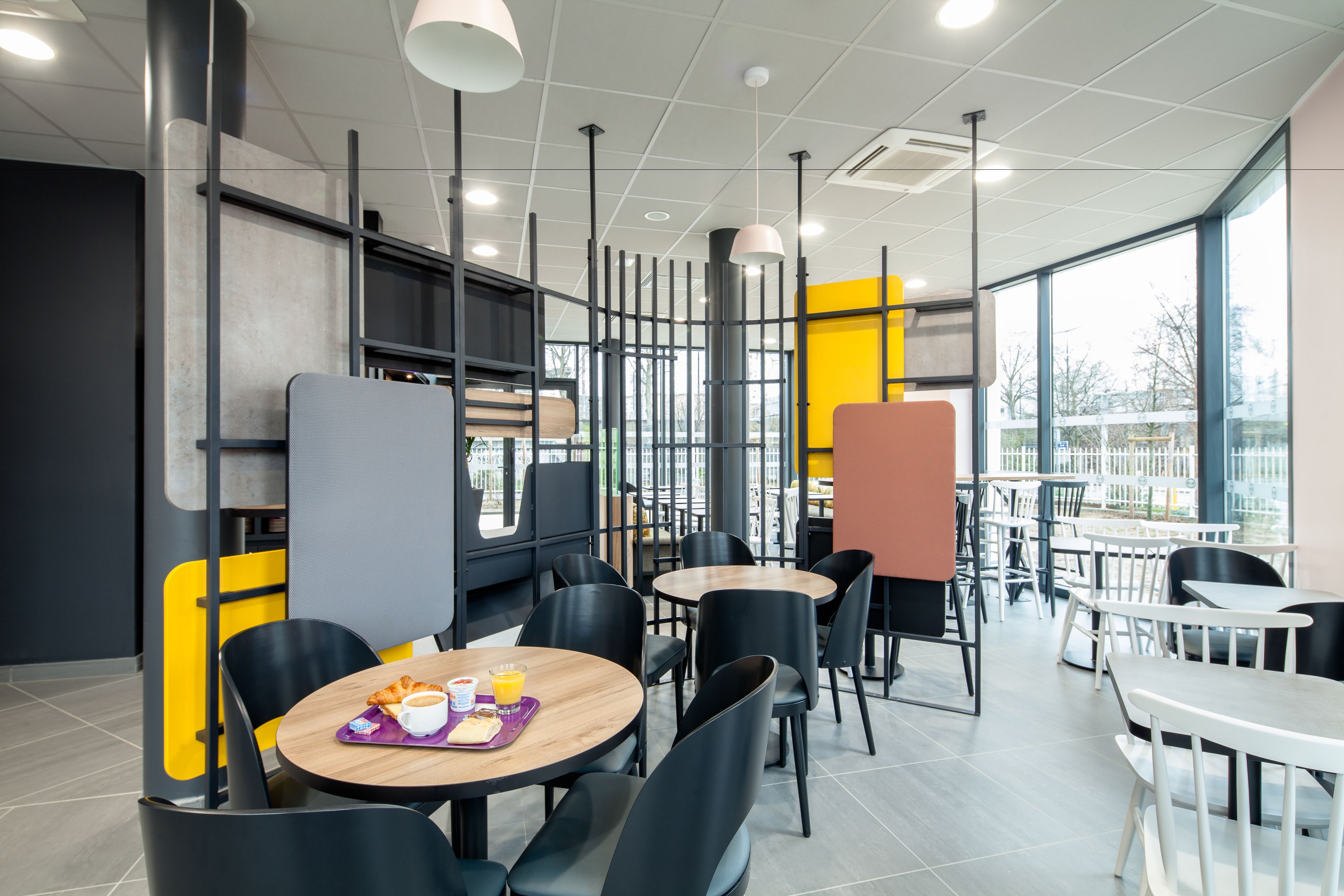 A modern cafe with round tables and black chairs, featuring a bright yellow accent wall and large windows.