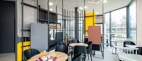 A modern cafe with round tables and black chairs, featuring a bright yellow accent wall and large windows.