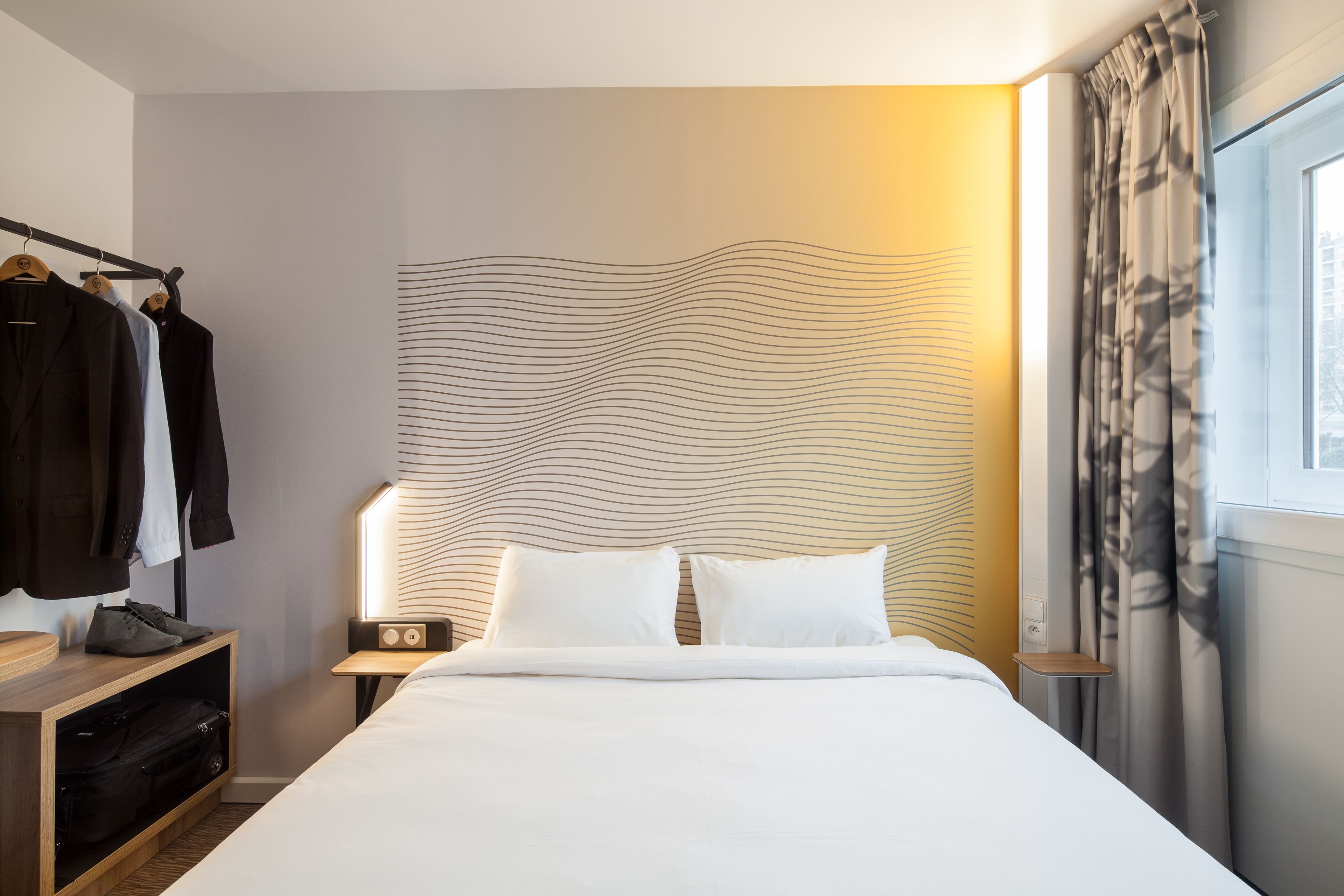 Una habitación de hotel con una cama bien hecha, una mesita de noche con lámpara, un armario con ropa colgada y una ventana con cortinas.