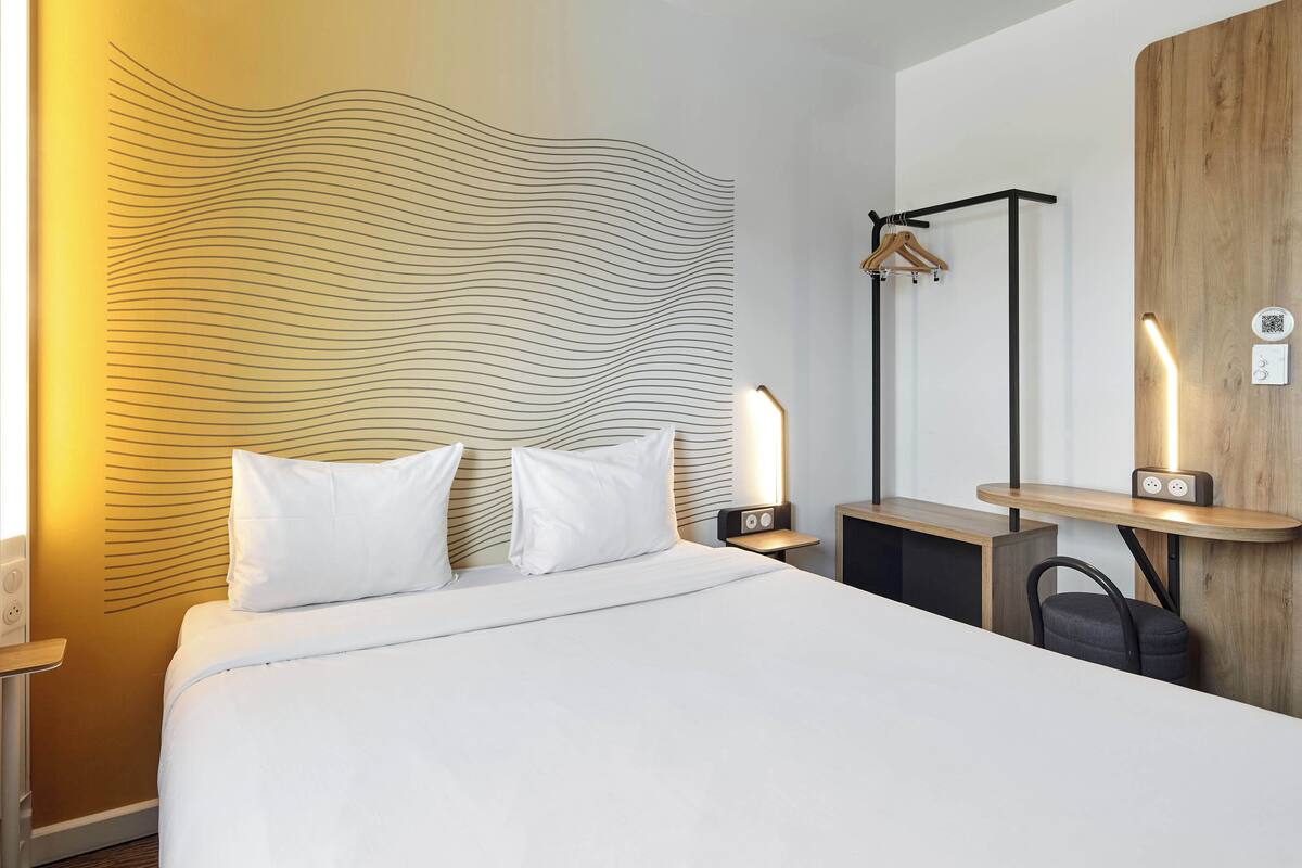 A modern hotel room with a large bed, a wooden headboard, a bedside table with a lamp, a wooden sideboard, and a hanging coat rack.