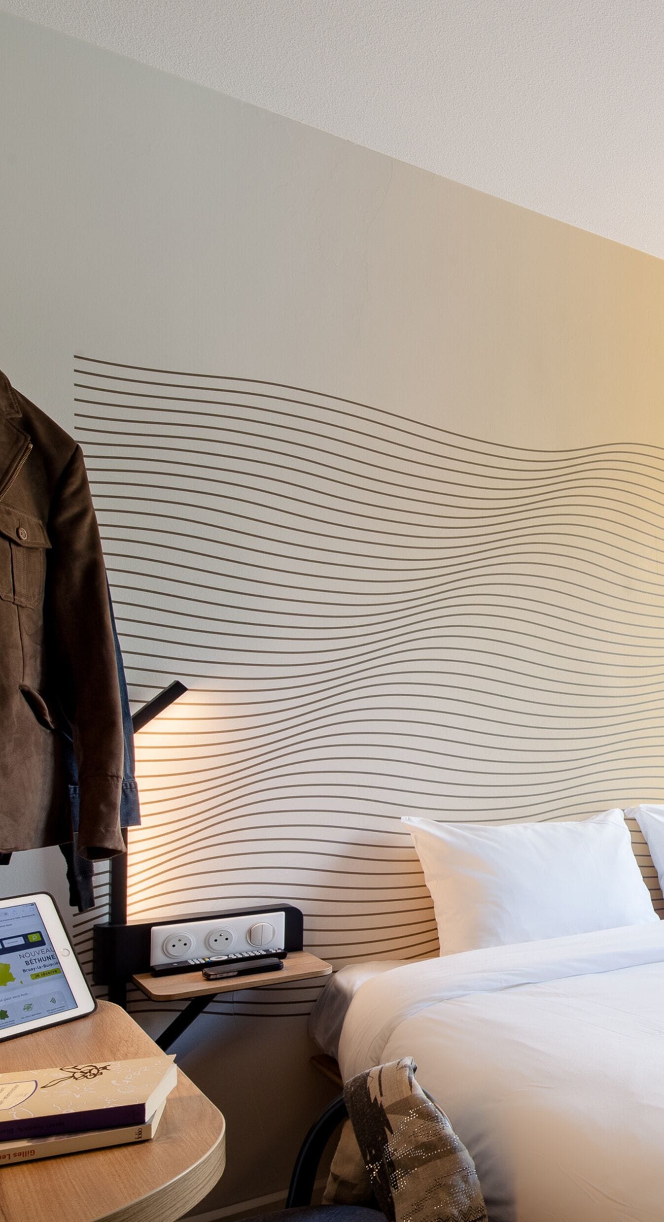 A hotel room with a bed, a bedside table with a lamp, a coat rack, and a wall with a wavy pattern.