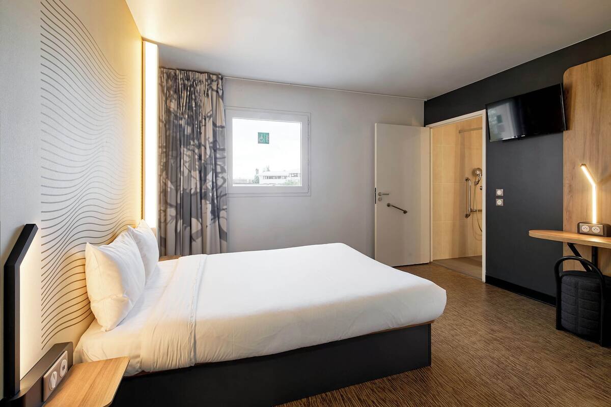 A modern hotel room with a large bed, a bedside table, a TV, and a window with curtains.