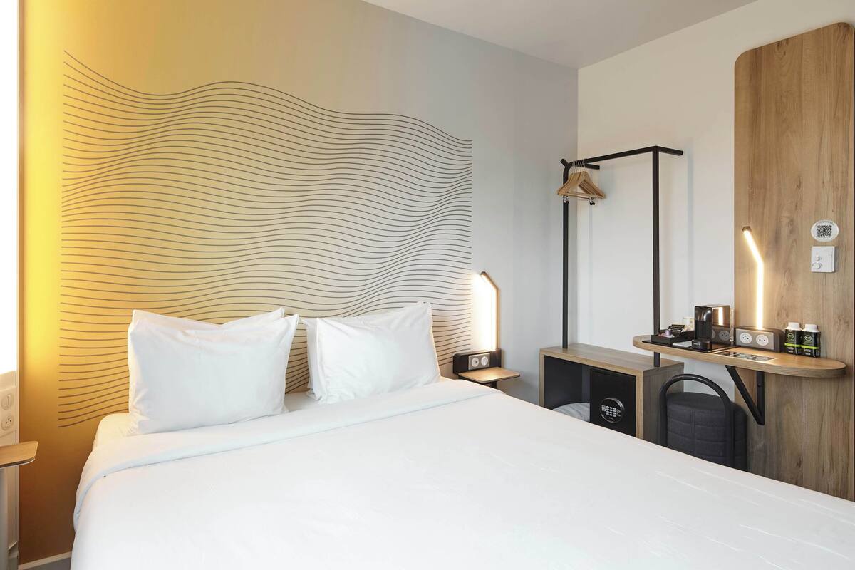 A modern hotel room with a large bed, a bedside table with a lamp, a wooden desk with a chair, and a wall with a wavy pattern.