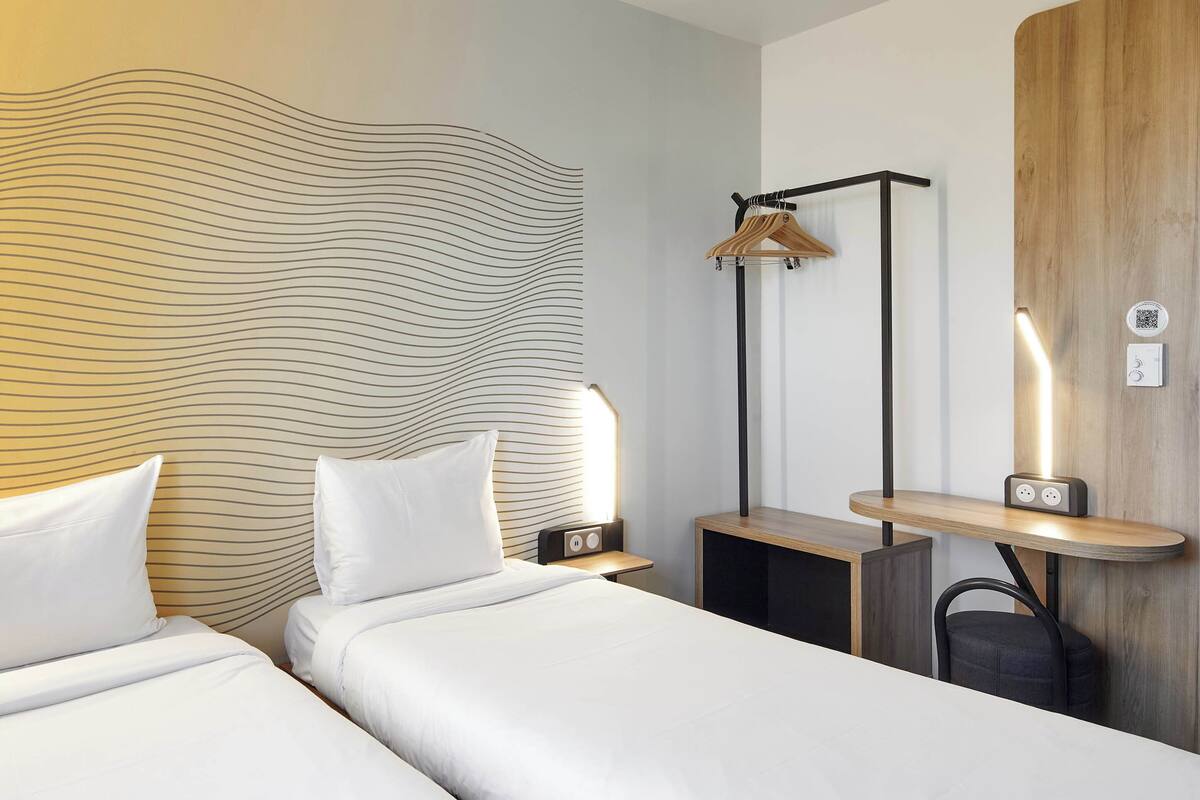 A modern hotel room with two beds, a wooden headboard, a hanging light, a bedside table, and a small shelf with a clock.