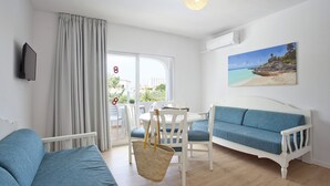 Una habitación con un sofá blanco, un sofá azul, una mesa de comedor con sillas, un ventanal con cortinas y un cuadro de playa en la pared.