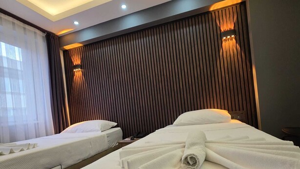 A hotel room with two beds, a large window with sheer curtains, and a wooden panel wall with recessed lighting. - Gizem Pansiyon (Çanakkale)