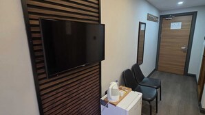 A hotel room with a flat-screen TV mounted on a wooden panel wall, a white minibar, two black chairs, and a wooden door with a sign. - Gizem Pansiyon (Çanakkale)