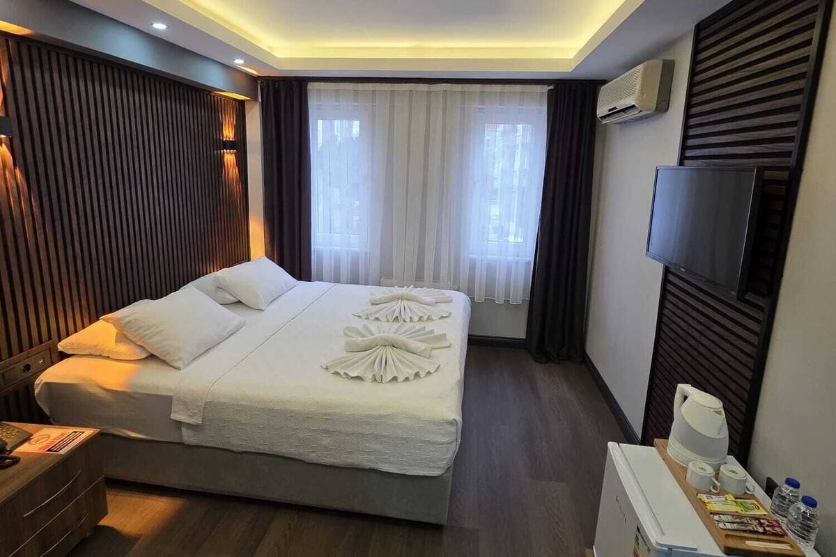 A hotel room with a large bed, a desk with a chair, a flat-screen TV, and an air conditioning unit.