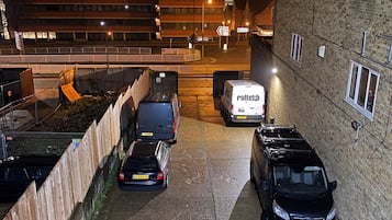 Un aparcamiento con varios coches y una furgoneta blanca, rodeado de edificios y una valla.
