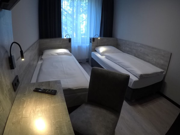 A small hotel room with two single beds, a desk with a chair, and a window with curtains. - Urban Home Self Check-In Hotel (Hamburg)