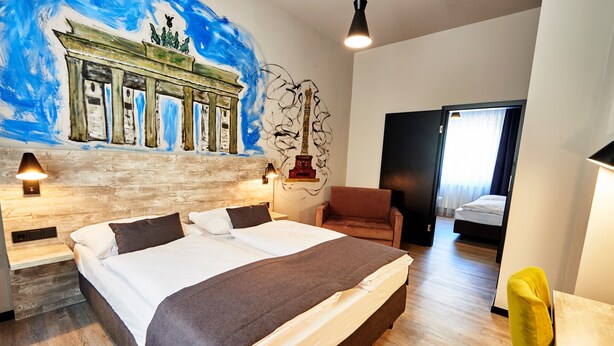 A hotel room with a large bed, a wooden headboard, a mural of a classical building, a statue, and a door leading to another room. - Urban Home Self Check-In Hotel (Hamburg)
