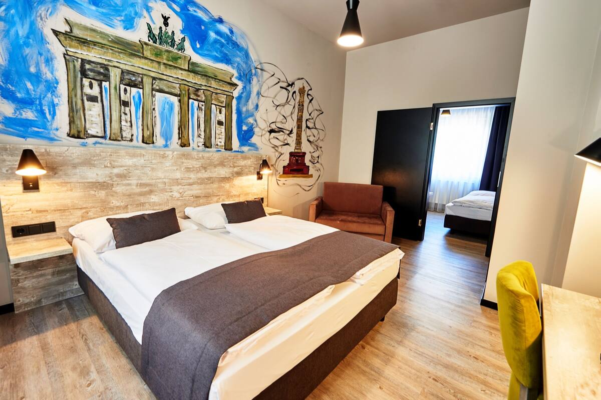 A hotel room with a large bed, a wooden headboard, a mural of a classical building, a statue, and a door leading to another room.
