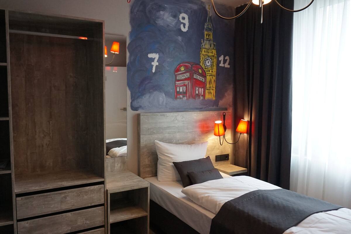 A hotel room with a bed, a wardrobe, a headboard with a painting, and a nightstand with a lamp.