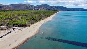 En kustnära strand med kristallklart turkost vatten, en sandstrand och ett skogsområde där bilar är parkerade.