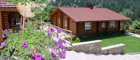 Una cabaña de madera con techo rojo, rodeada por un muro de piedra y una escalera de piedra que lleva a una terraza de madera.