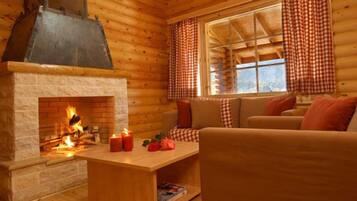 A cozy cabin interior with a fireplace, wooden walls, a sofa, and a window with checkered curtains.