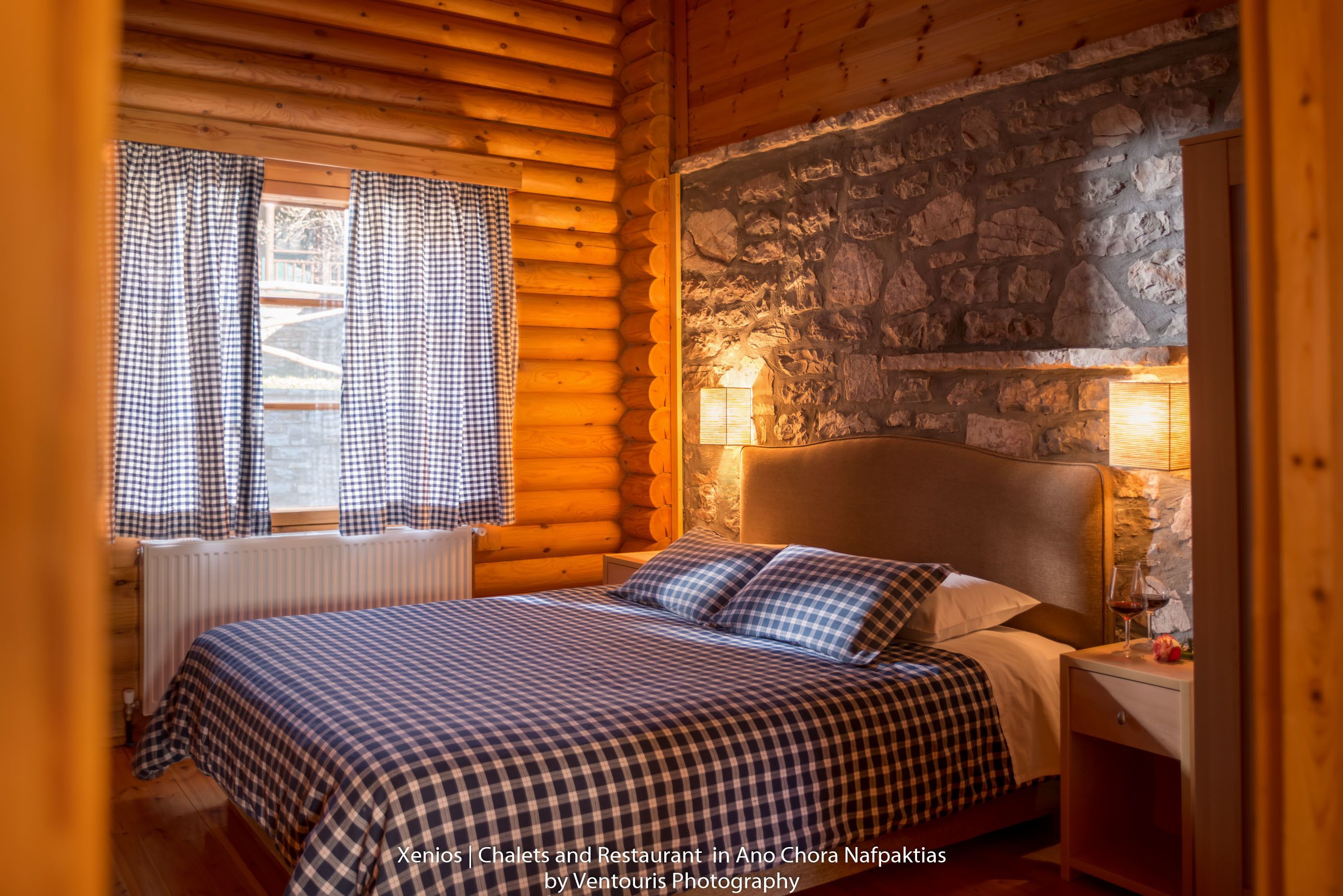 Quarto de cabana de madeira com cama, cortinas xadrez, parede de pedra e mesa de cabeceira com um copo de vinho.