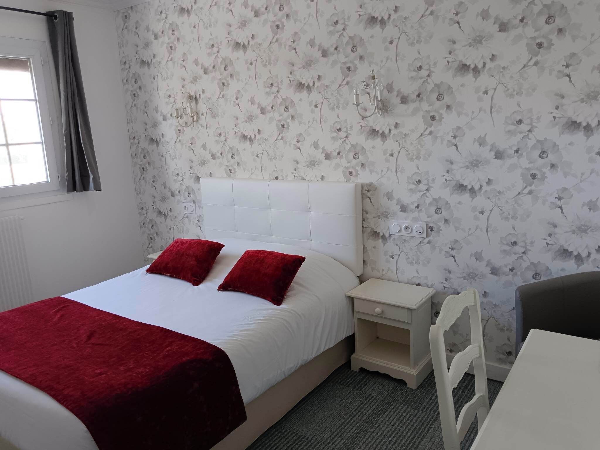 Un dormitorio con una cama blanca, almohadas rojas, una mesita de noche blanca, una silla blanca y papel tapiz floral.