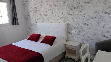 Un dormitorio con una cama blanca, almohadas rojas, una mesita de noche blanca, una silla blanca y papel tapiz floral.