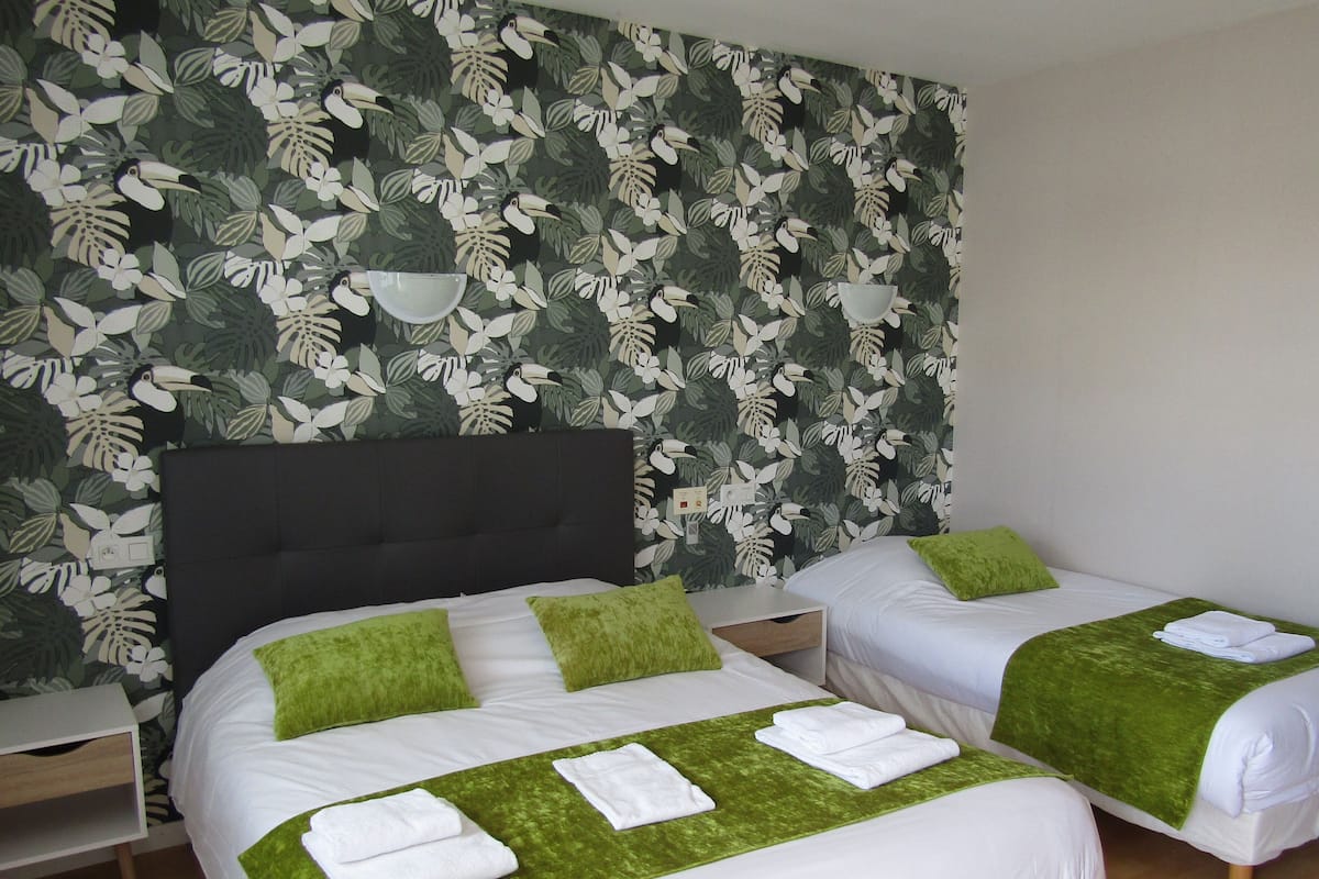 Habitación de hotel con dos camas, cada una con almohadas verdes y ropa de cama blanca. La habitación tiene un ventanal grande y una pared con estampado de hojas tropicales.
