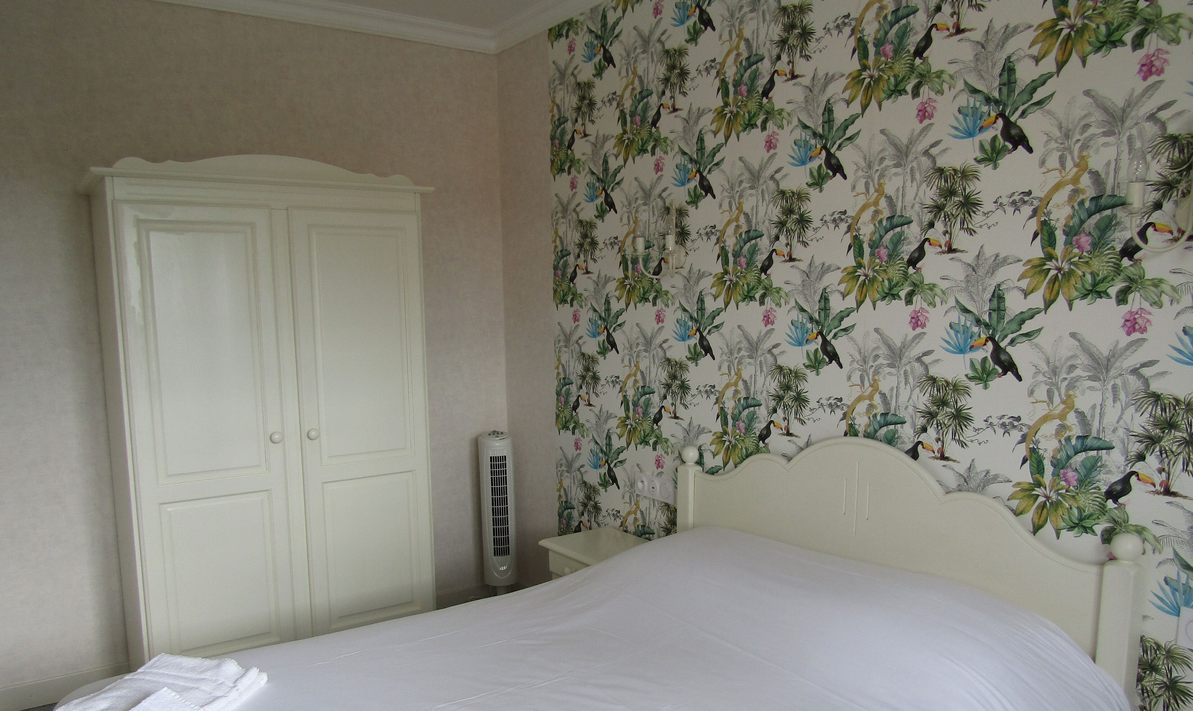 A bedroom with a large white wardrobe, a bed with white bedding, and a wall with a tropical wallpaper.