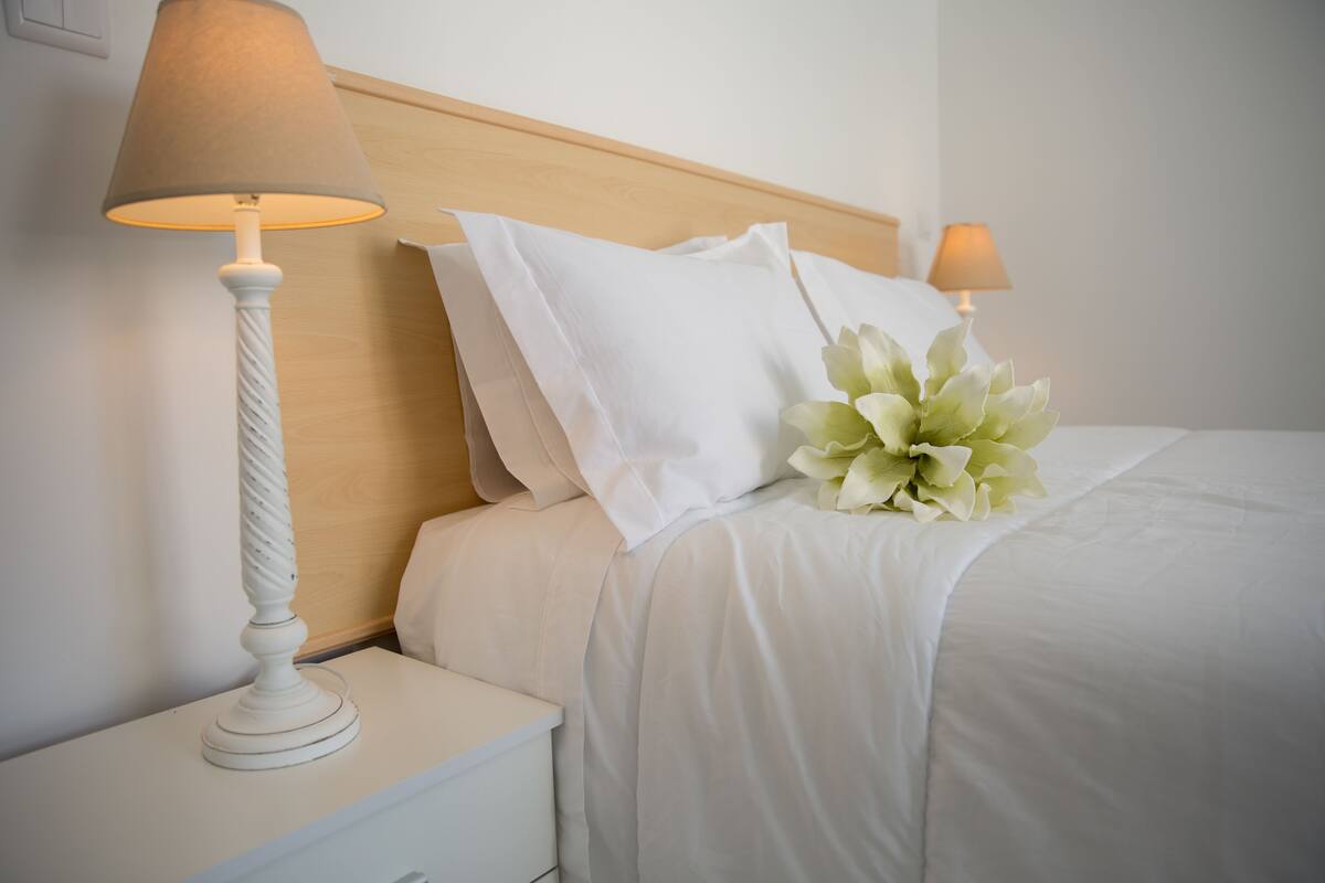 A neatly made bed with white linens and a bouquet of white flowers, a white bedside table with a lamp, and a wooden headboard.