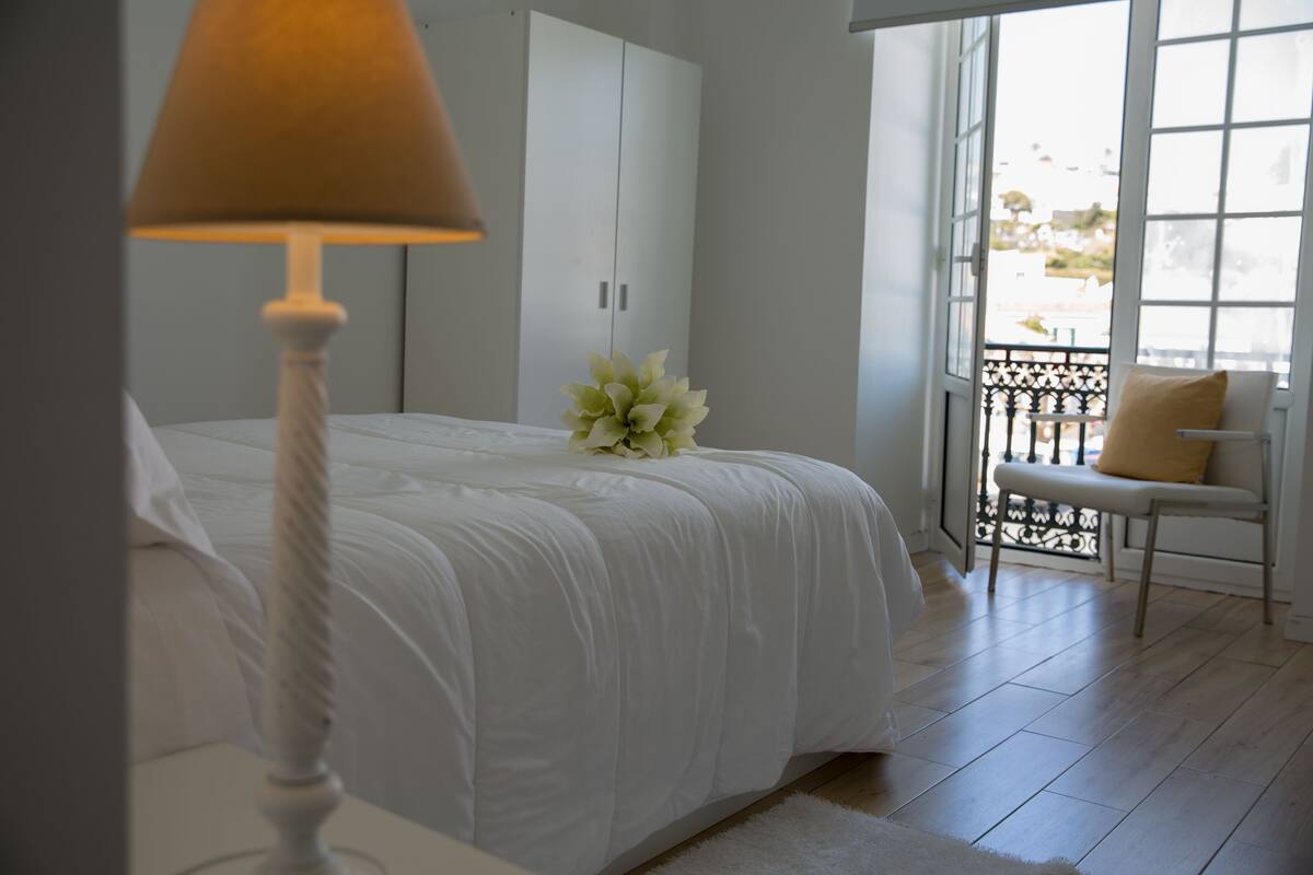 A bedroom with a bed, a white lamp, a bench, and a view of the outside through a window.