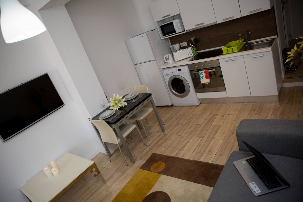 A modern kitchen with a washing machine, microwave, and sink. There is a dining area with a table and chairs, and a sofa with a laptop on it.