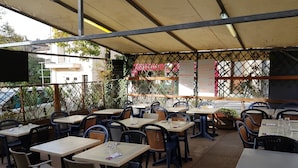 Ein Straßencafé mit Tischen und Stühlen, das von einem Sonnensegel überdacht und von Grünpflanzen umgeben ist.