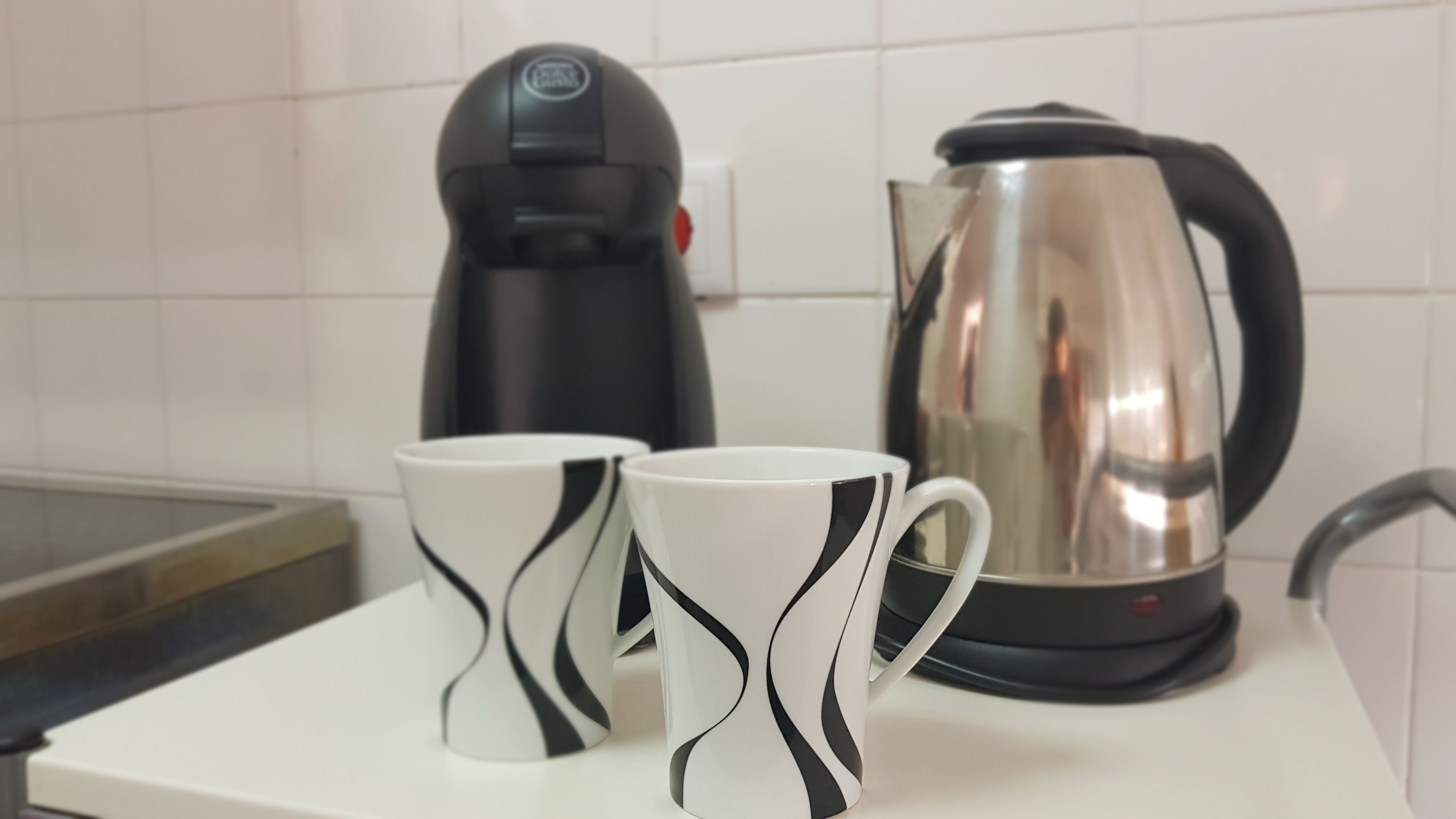Two white mugs with black wavy patterns on a white countertop. A black coffee maker and a stainless steel electric kettle are visible in the background.