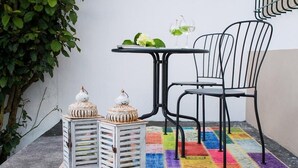 Uma pequena área externa com mesa e duas cadeiras, um tapete colorido e dois lanternas brancas com detalhes decorativos.
