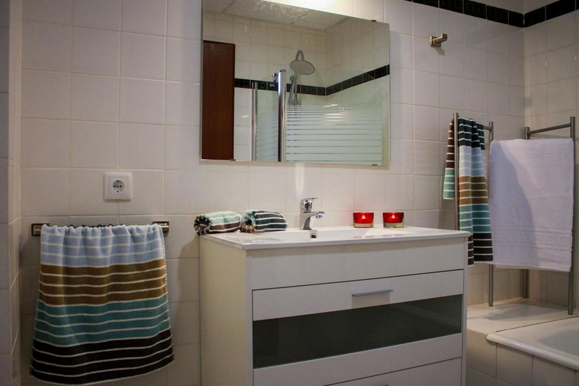 A bathroom with a white sink, mirror, and a shower. There are striped towels on the wall and a red candle on the sink.