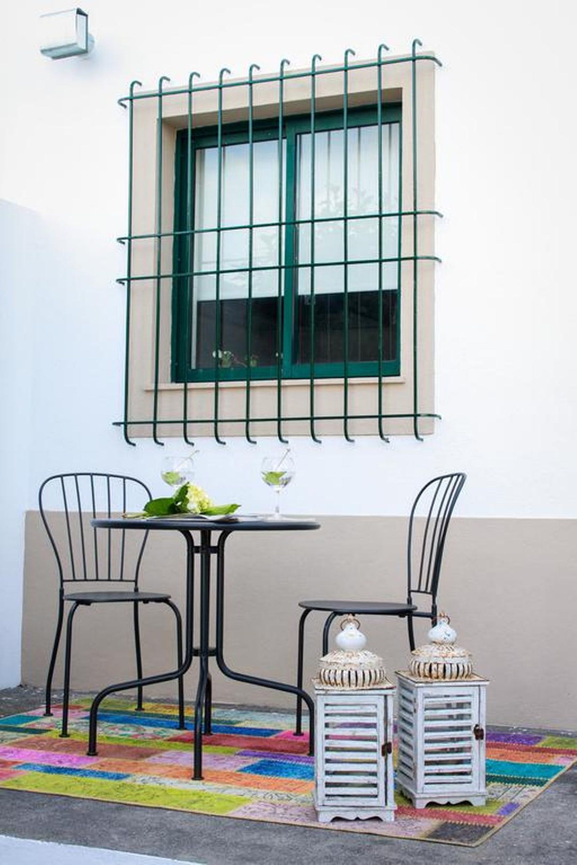 A small outdoor seating area with a table and two chairs, a window with a green frame, and two white lanterns on a colorful rug.
