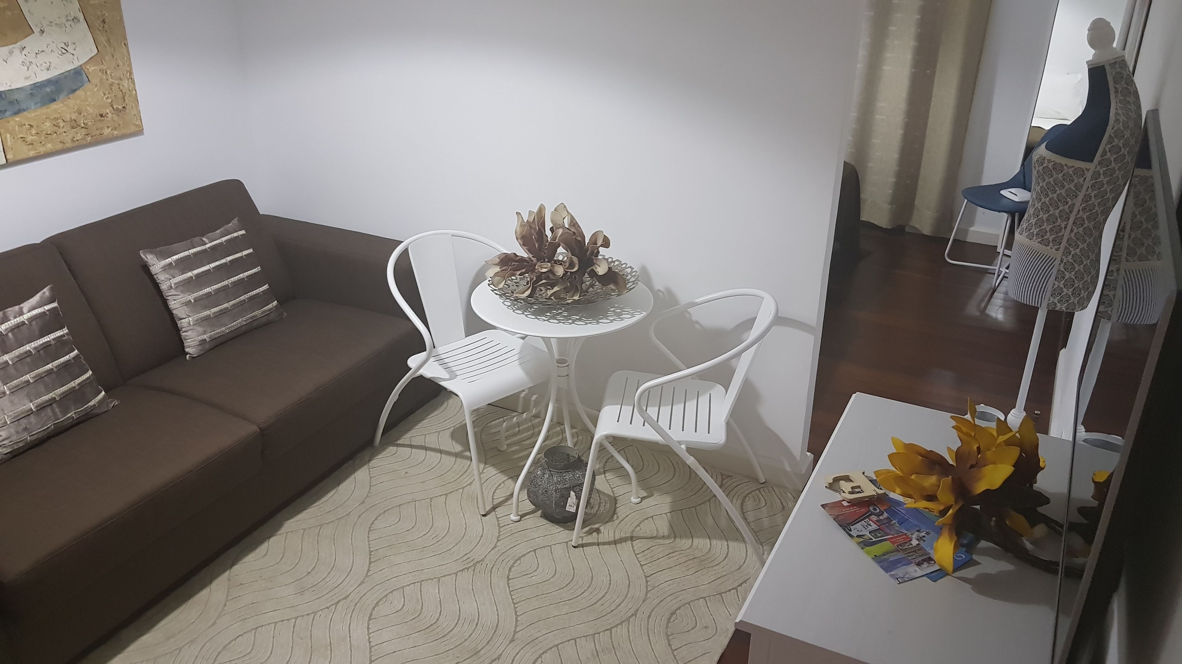 A living room with a brown sofa, white chairs, a small round table with a decorative bowl, and a small table with yellow flowers.